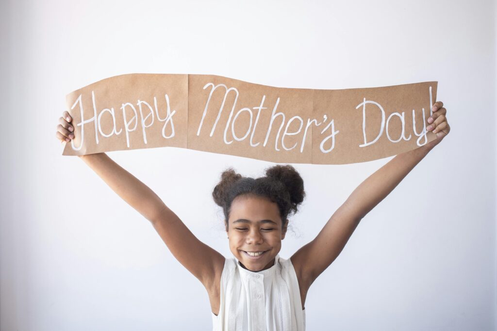 Smiling child holding a 'Happy Mother's Day' sign, celebrating indoors.